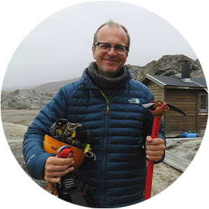 Justin Francis OBE, a man standing on rocky terrain, on a visit to Spitsbergen in the Norwegian Arctic, wearing glasses and cold weather clothes, smiling at the camera and carrying outdoor gear including a helmet and pick axe