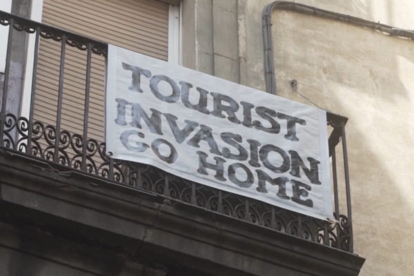 Film still of sign saying 'tourist invasion go home' hanging on balcony railings